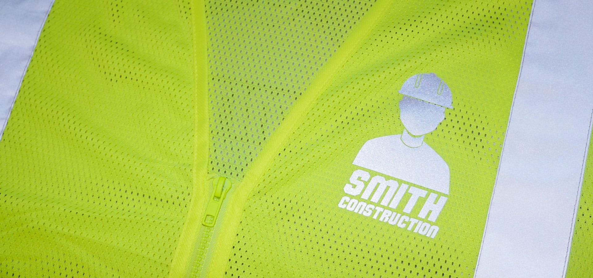 A yellow safety vest with a construction company logo pressed on it in Reflection Protection™. It has light shown on it to show how reflective it is.