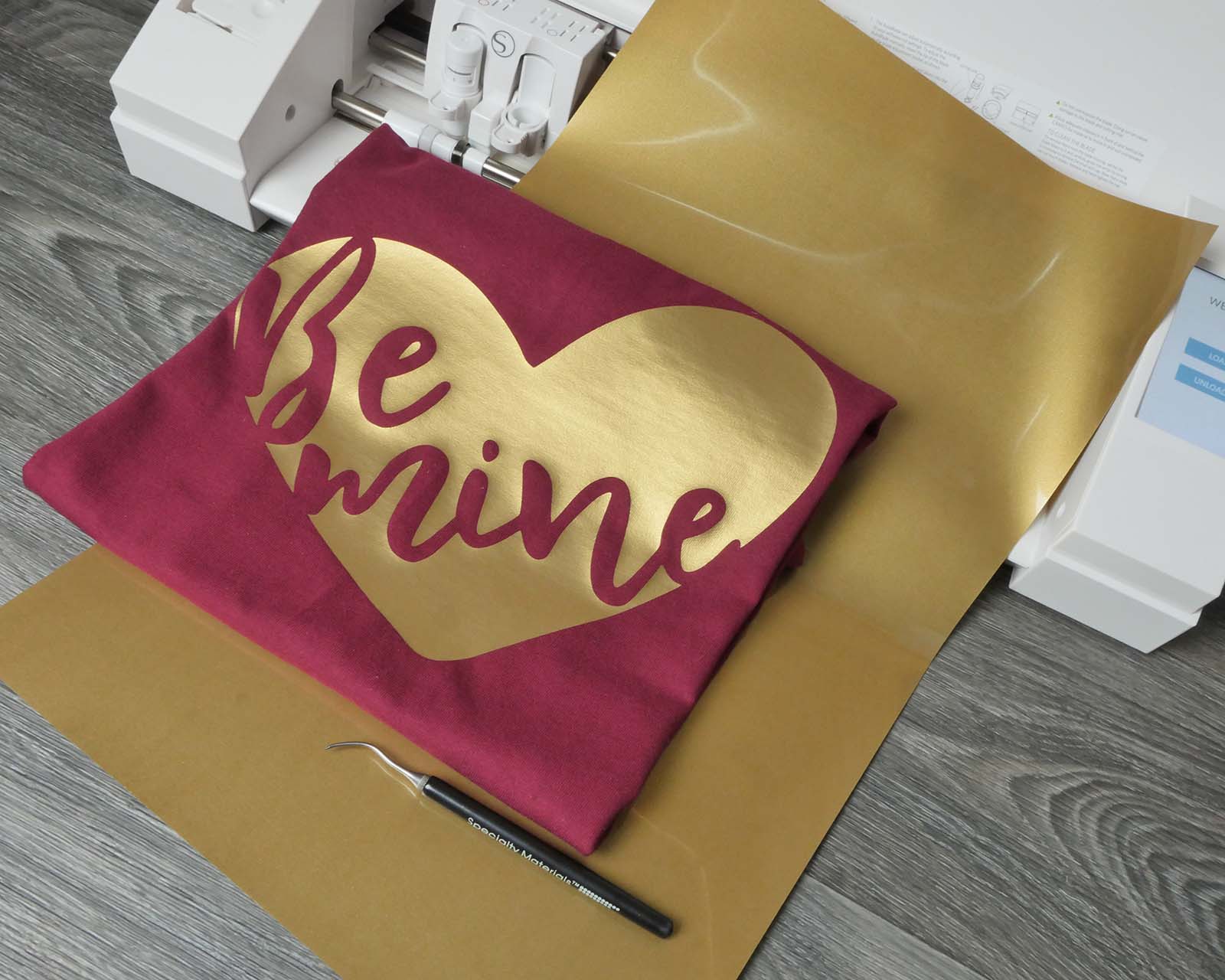A shirt with a design that read "Be Mine" in a heart made with Dark Gold ThermoFlex Plus laying on top of a Silhouette Cameo 3.